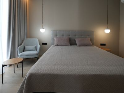 Elegantes Schlafzimmer mit grau gepolstertem Bett, zwei modernen Pendelleuchten und einem bequemen Sessel. Helle Vorhänge und Holzbodenoptik schaffen eine ruhige Atmosphäre.