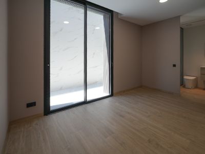 Geräumiges Zimmer mit Holzoptikfliesen und hellen Wänden. Große schwarze Glasschiebetüren führen zu einem Außenbereich mit weißen Marmorfliesen. Integriertes Badezimmer sichtbar.