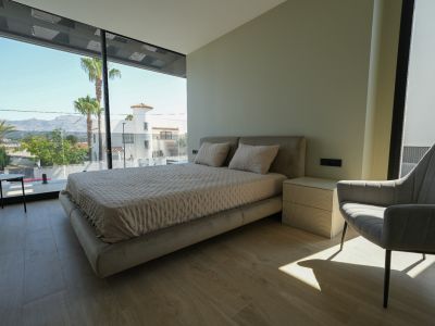 Hauptschlafzimmer mit gepolstertem Bett, geripptem Nachttisch und raumhohen Fenstern mit Blick auf Palmen und Berge.