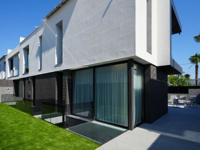 Moderne Außenansicht der Villa mit kontrastierender weiß-grauer und dunkler Fassade, großen Glasfronten, Kunstrasenfläche und gefliester Terrasse mit Sitzgelegenheiten.