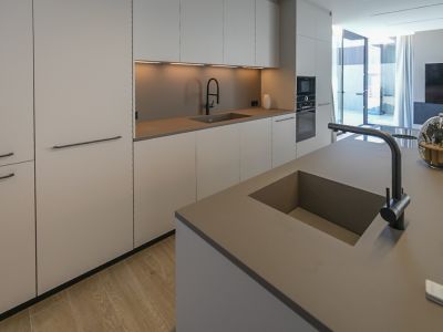 Moderne Küche mit Kücheninsel und integriertem Doppelwaschbecken. Minimalistische Schränke in neutralen Tönen, Einbaugeräte und heller Holzfußboden, mit Zugang zum Außenbereich.