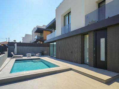 Moderner Villa-Außenbereich mit rechteckigem Pool und integrierten Stufen, umgeben von hellem Pflaster. Dunkle Lamellenverkleidung der Erdgeschossfassade und markante Eingangstür.
