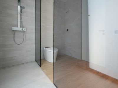 Modernes Badezimmer mit ebenerdiger Dusche, hellgrauen Fliesen und verchromter Thermostatarmatur. Eine Glaswand trennt die Dusche vom Smart-WC-Bereich mit Holzoptik-Fliesenboden.