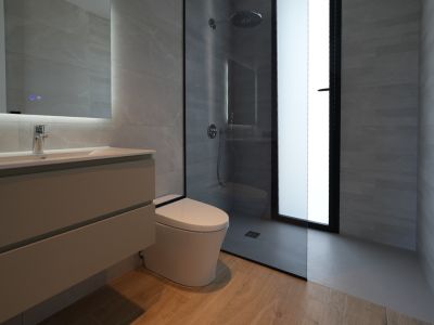 Modernes Badezimmer mit schwebendem Waschtisch, beleuchtetem Spiegel, wandhängendem WC und ebenerdiger Dusche mit Regenduschkopf, ergänzt durch graue Fliesen und Holzbodenoptik.
