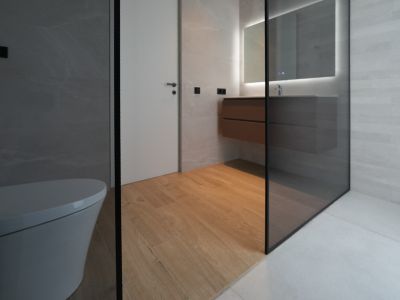 Modernes Badezimmer mit wandhängendem WC, schwebendem Holzwaschtisch, beleuchtetem Spiegel und Glasduschabtrennung. Holzbodenfliesen ergänzen die grauen Wand- und Duschfliesen.