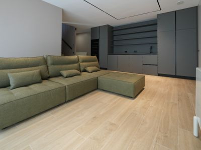 Olivgrünes Modulsofa in offenem Wohnbereich mit hellem Holzoptik-Fliesenboden. Im Hintergrund dunkle Einbauschränke mit Spüle und Regalen.