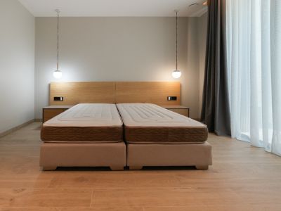 Schlafzimmer mit Doppelbett aus zwei Polsterbetten, hellem Holzkopfteil mit integrierten Nachttischen und zwei modernen Pendelleuchten. Großes Fenster mit Vorhängen.