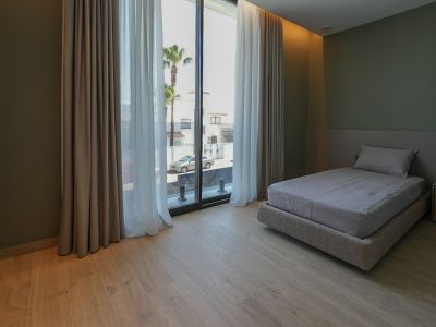 Schlafzimmer mit Einzelbett, hellem Holzfußboden und olivgrünen Wänden. Große Fenster mit Doppelvorhängen bieten Blick auf die Straße und Palmen.