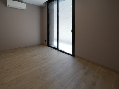 Unmöblierter Raum mit hellem Holzfliesenboden und taupefarbenen Wänden. Eine schwarze Glasschiebetür und eine Wandklimaanlage sind sichtbar.
