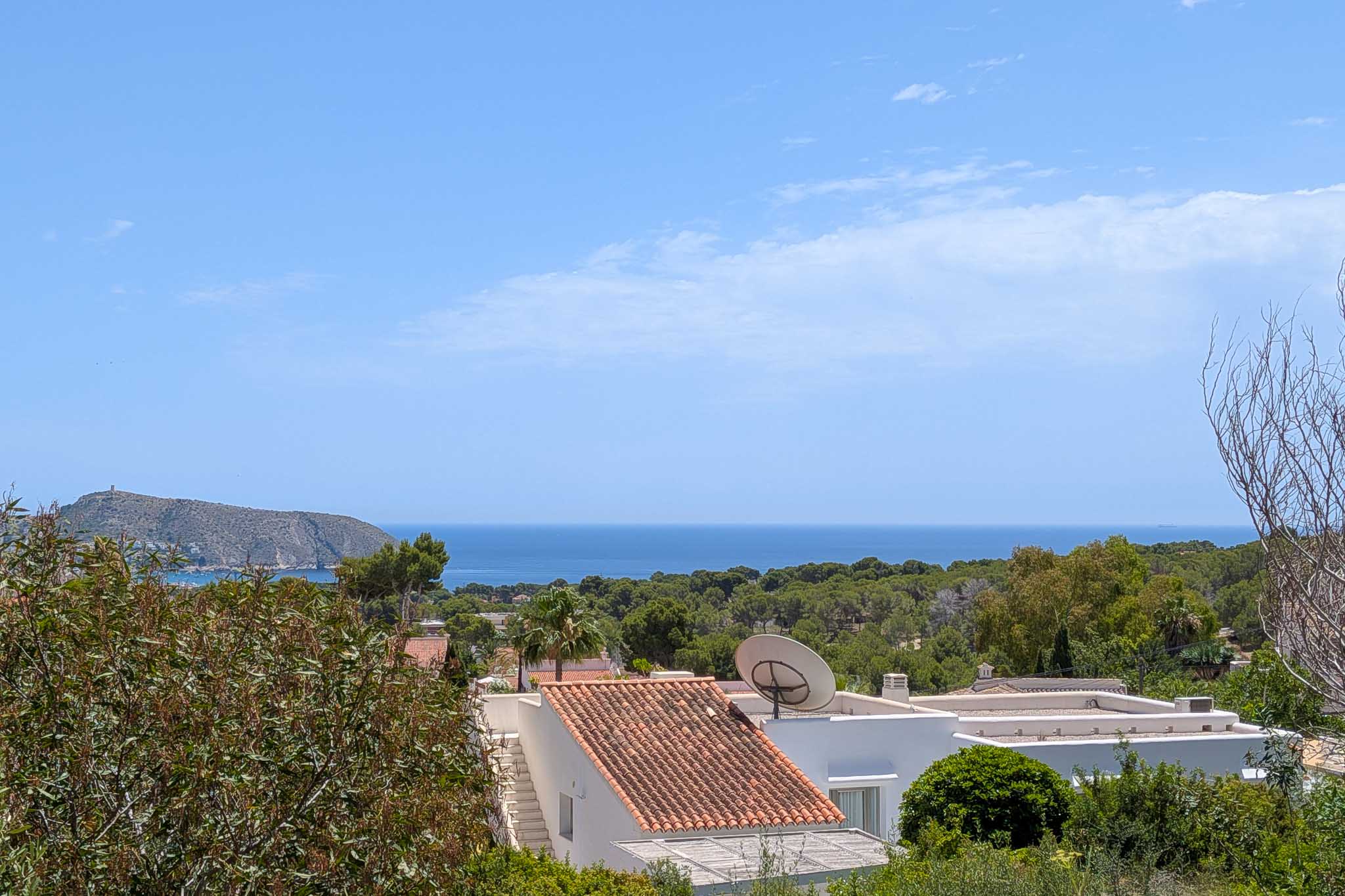 Erhöhter Meerblick auf das Mittelmeer, mit traditionellen Ziegeldächern und üppiger Vegetation im Vordergrund. Eine felsige Landzunge mit Turm ist in der Ferne unter blauem Himmel sichtbar.