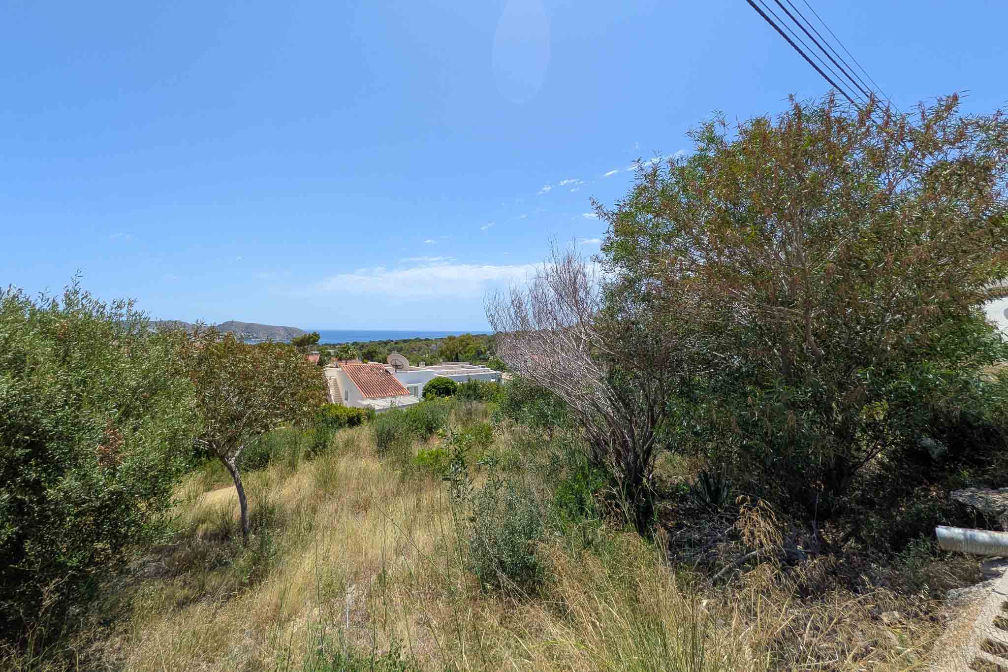 Naturbelassenes Grundstück mit trockener Mittelmeervegetation, mit Fernblick auf Meer und Berge unter blauem Himmel. Leicht abfallendes Gelände.