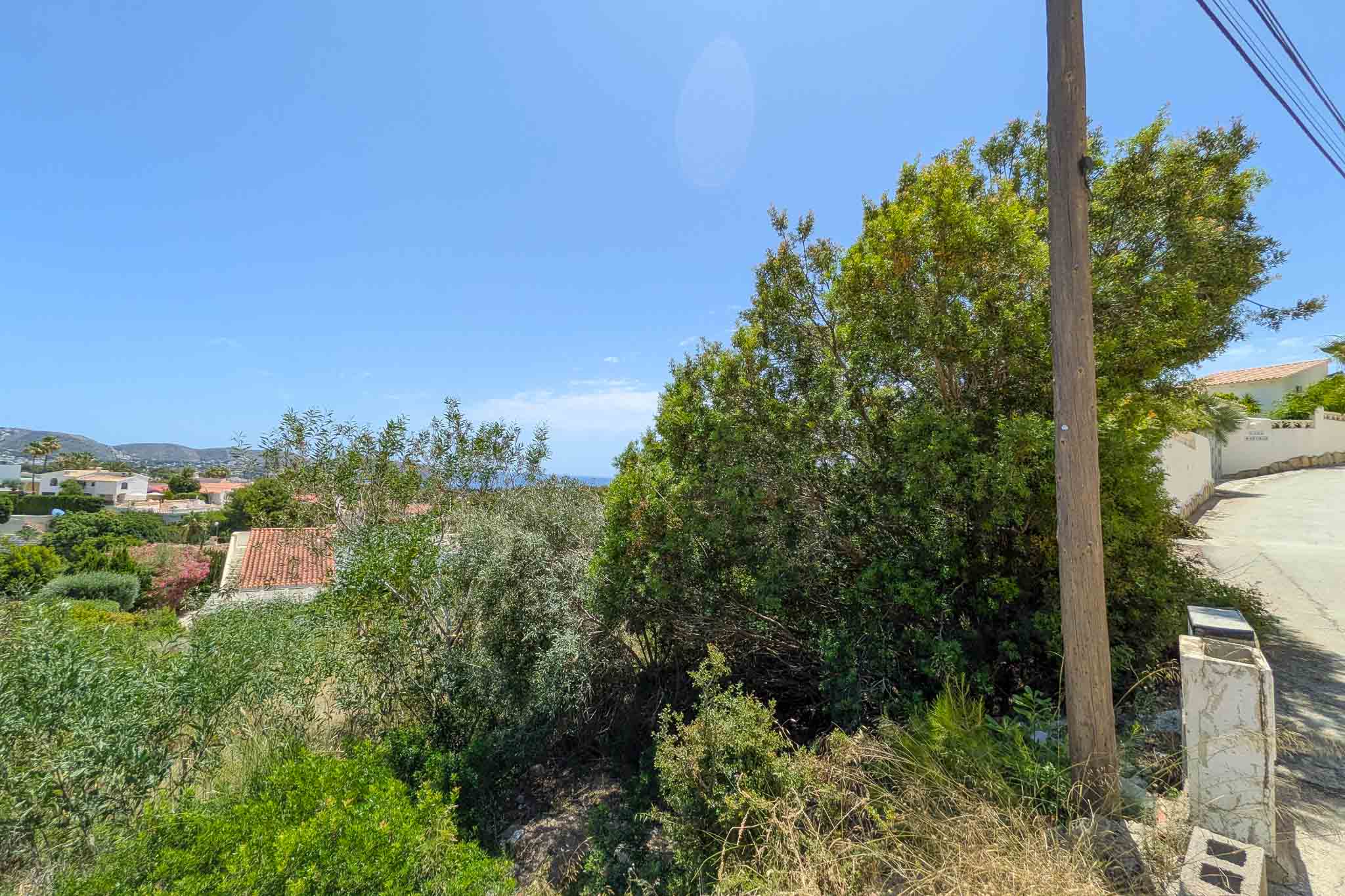 Unbebautes Grundstück mit dichter mediterraner Vegetation, Strommast und gepflastertem Weg. Bietet Fernblick auf Dächer, Hügel und blauen Himmel mit Meeresglitzern.