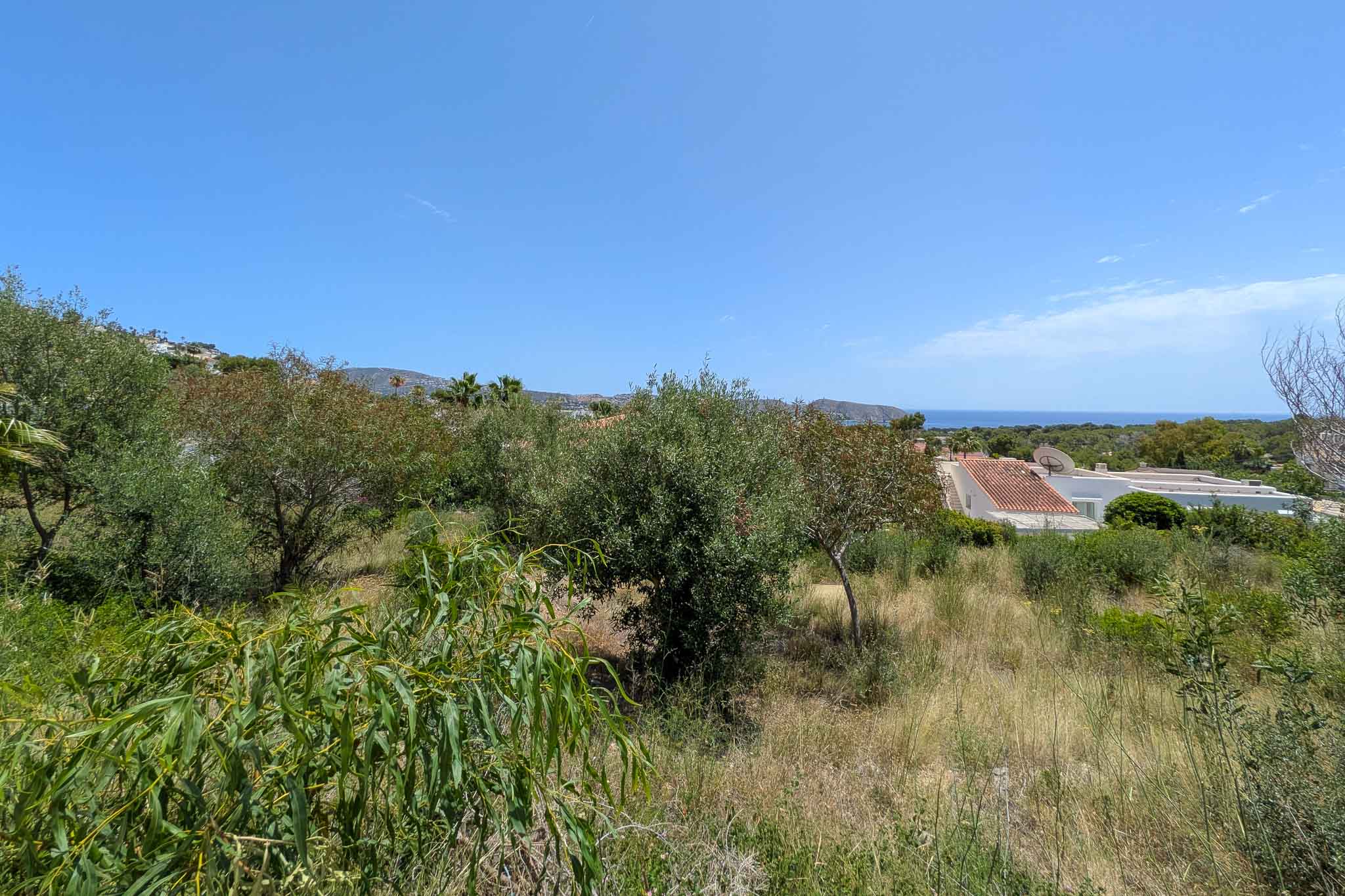 Unbebautes Grundstück mit natürlicher mediterraner Vegetation, das partielle Meerblicke, umrahmt von Wohndächern und fernen Hügeln unter klarem blauem Himmel, bietet.