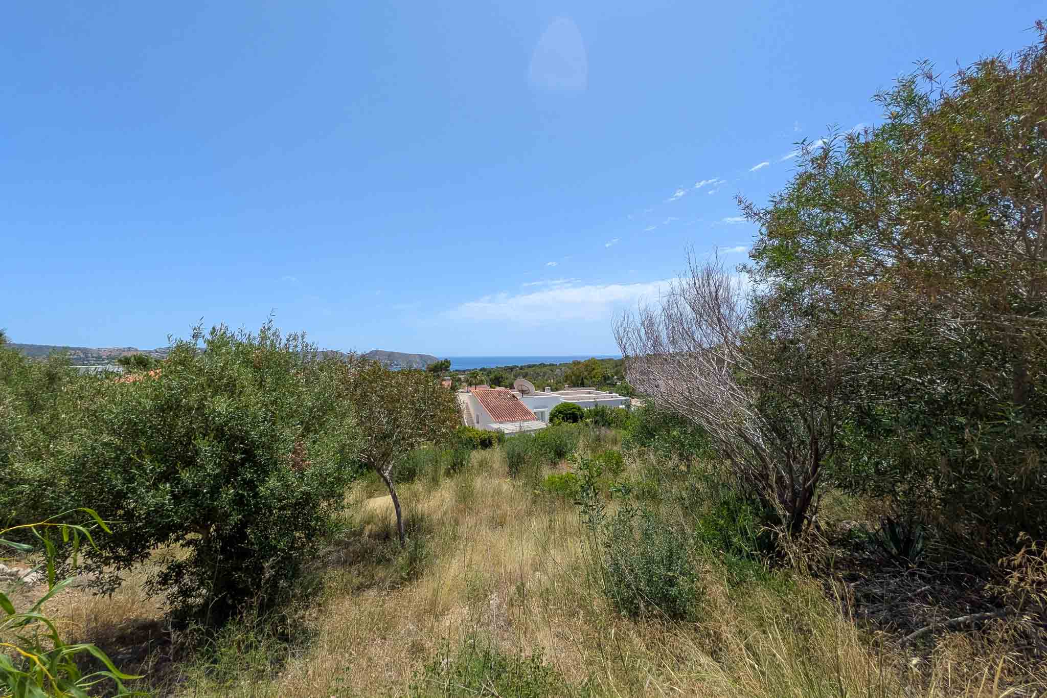 Weitläufiges Grundstück mit natürlicher Vegetation und fernem Meerblick unter klarem blauem Himmel. Wohndächer sind im Mittelgrund sichtbar, umrahmt von grünen und trockenen Sträuchern.