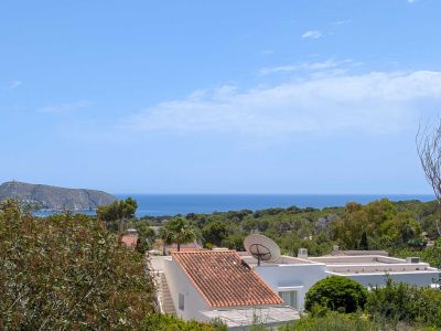 Erhöhter Meerblick auf das Mittelmeer, mit traditionellen Ziegeldächern und üppiger Vegetation im Vordergrund. Eine felsige Landzunge mit Turm ist in der Ferne unter blauem Himmel sichtbar.