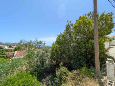 Unbebautes Grundstück mit dichter mediterraner Vegetation, Strommast und gepflastertem Weg. Bietet Fernblick auf Dächer, Hügel und blauen Himmel mit Meeresglitzern.