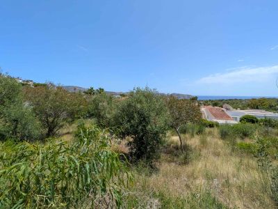 Unbebautes Grundstück mit natürlicher mediterraner Vegetation, das partielle Meerblicke, umrahmt von Wohndächern und fernen Hügeln unter klarem blauem Himmel, bietet.