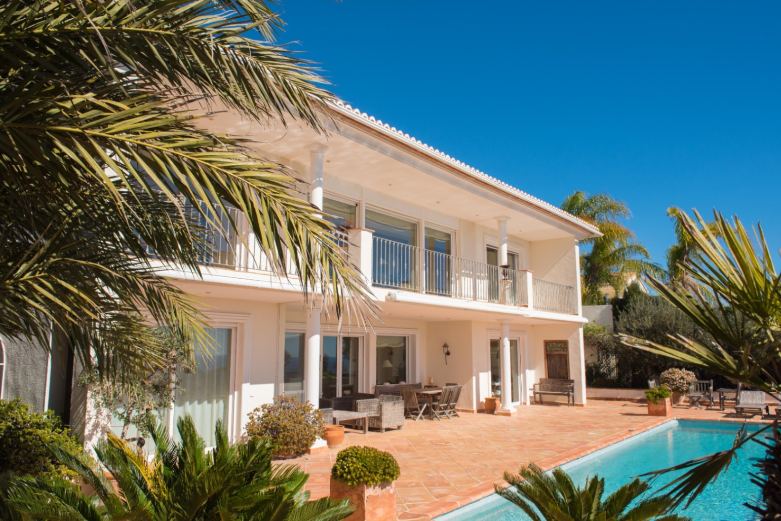 Außenansicht der zweistöckigen Villa mit Terrakotta-Terrasse, privatem Pool und oberem Balkon mit weißen Säulen und Metallgeländern.