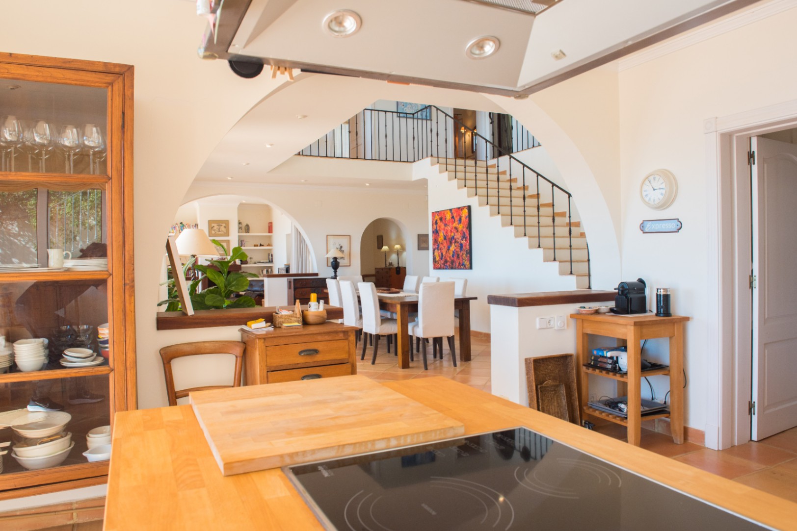 Blick von der Kücheninsel mit Holzoberfläche auf den Essbereich und die Treppe, akzentuiert durch mediterrane Rundbögen und helle Fliesen.