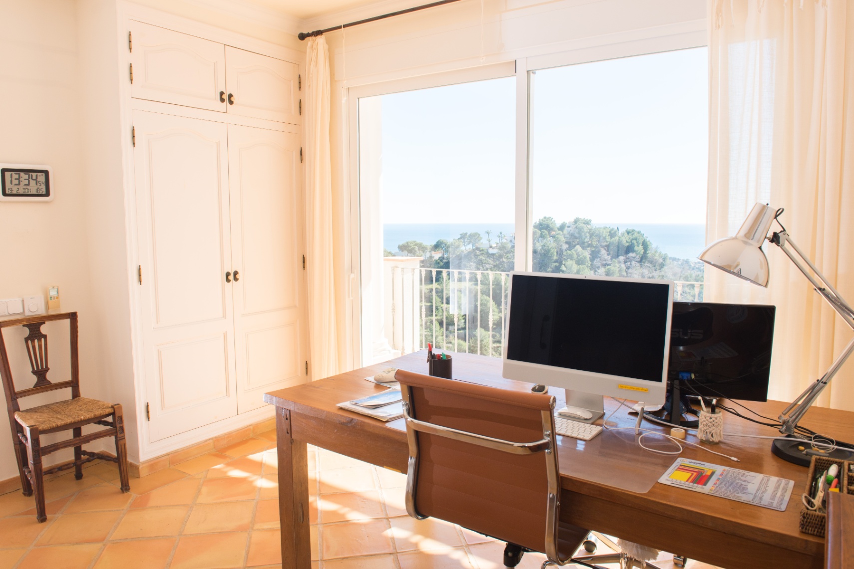 Helles Arbeitszimmer mit Holzschreibtisch, weißen Einbauschränken und bodentiefen Fenstern mit Panoramablick auf das Meer und die Küstenlandschaft.