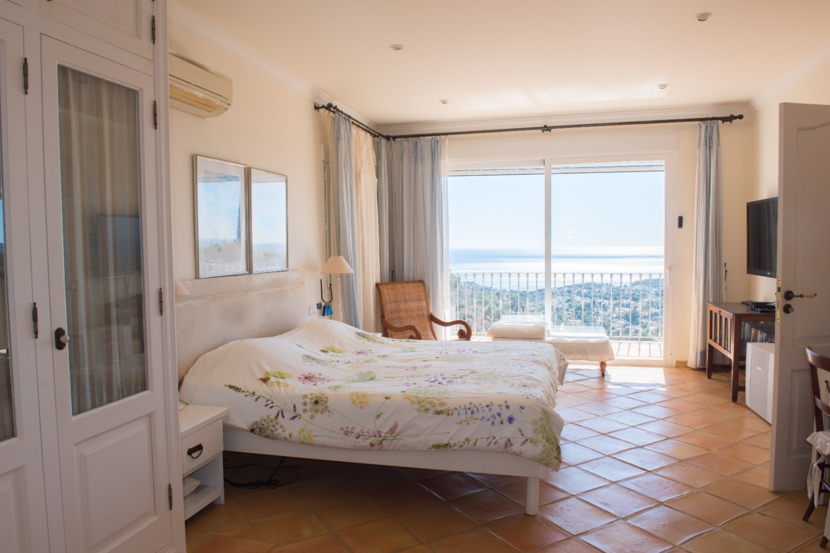 Helles Schlafzimmer mit Terrakotta-Fliesenboden, weißen Einbauschränken und Glasschiebetüren, die auf einen Balkon mit weitem Meerblick führen.