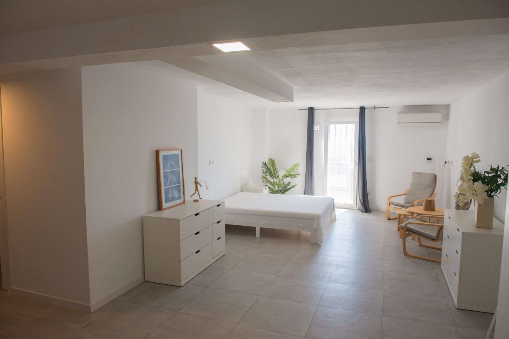 Minimalistisches Schlafzimmer in neutralen Tönen mit grauen Keramikböden, funktionalen weißen Möbeln, Klimaanlage und direktem Außenzugang.