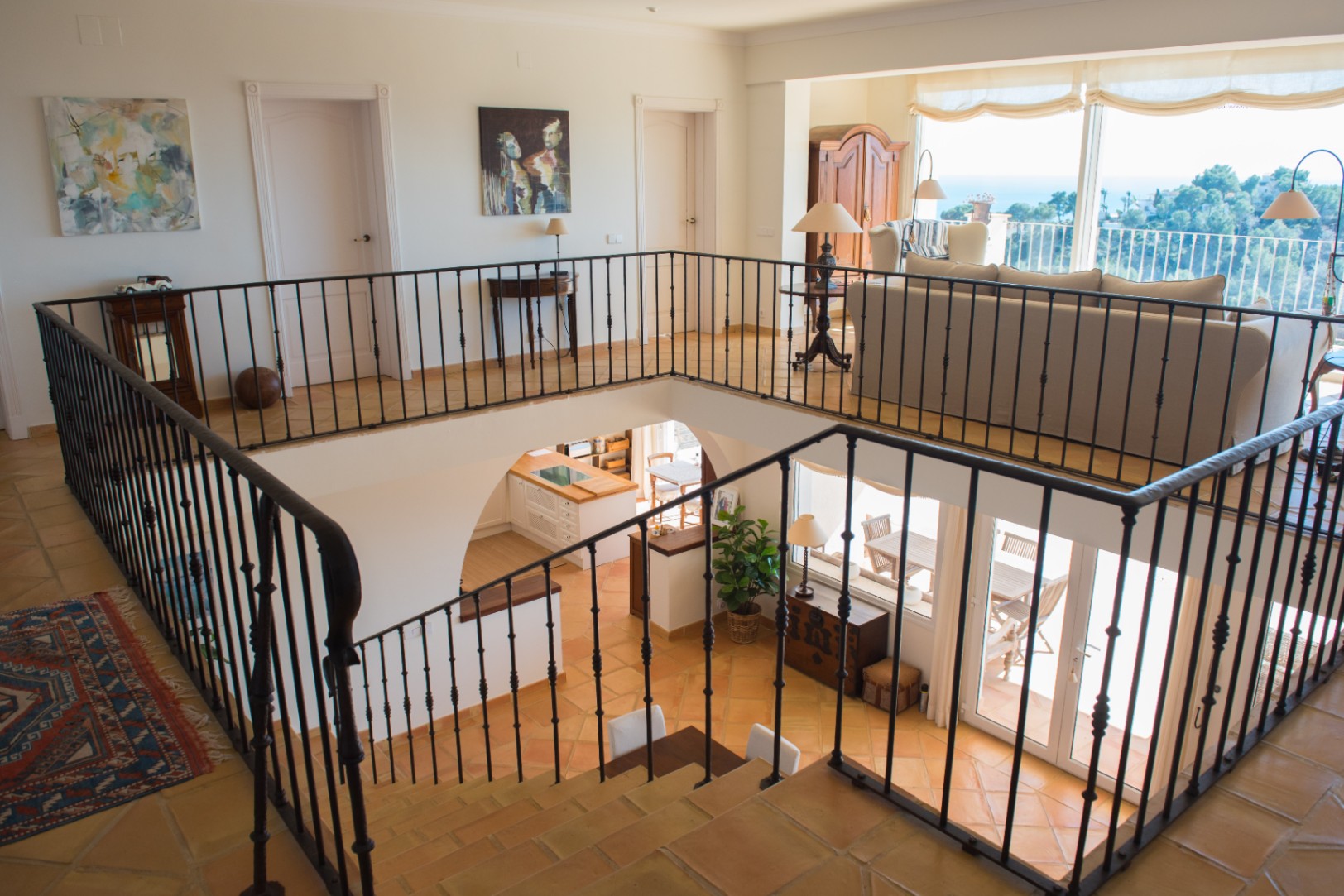 Obere Galerie mit schmiedeeisernem Geländer und Terrakottaboden, Blick auf den unteren Wohnbereich und Panoramafenster mit weitem Meerblick.
