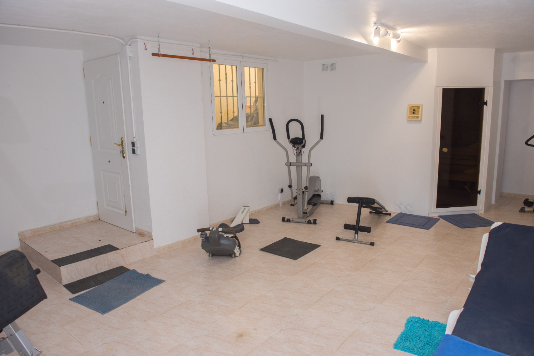 Privater Fitnessraum mit Kardiogeräten, Hantelbank und direktem Zugang zu einer Sauna. Der Raum ist mit hellen Fliesenböden ausgestattet.