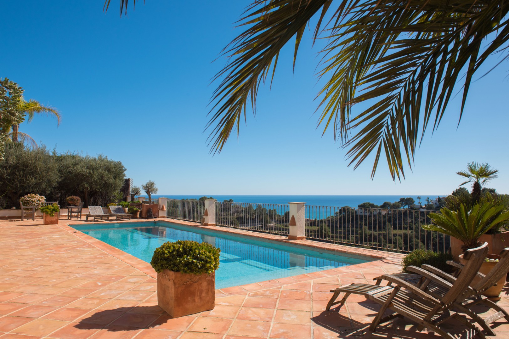 Terrakotta-Terrasse mit rechteckigem Pool, Holzliegen und Panoramablick auf das Mittelmeer, umrahmt von Palmen und mediterraner Vegetation.