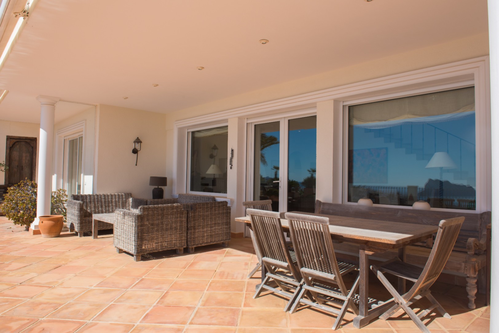 Überdachte Terrasse mit Terrakottaboden, Essbereich aus Holz und Rattan-Lounge-Möbeln vor großen Fensterfronten.