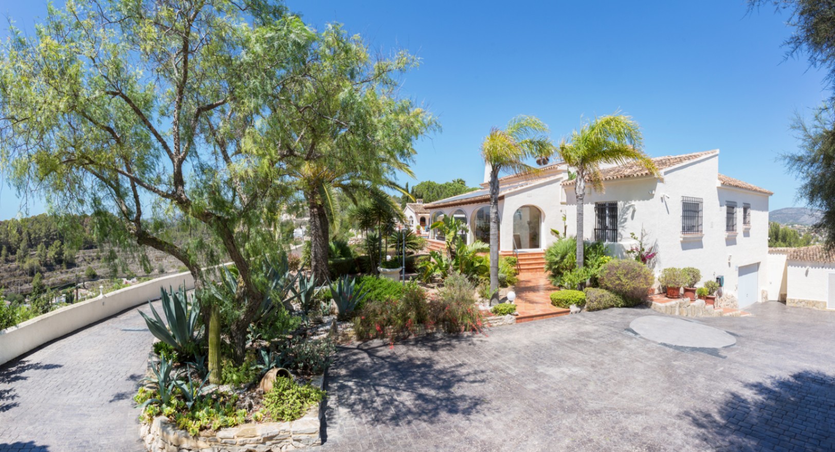 Außenansicht der mediterranen Villa mit gepflasterter Einfahrt, angelegtem Garten mit Agaven und Palmen sowie weißer Fassade mit Rundbogenfenstern.