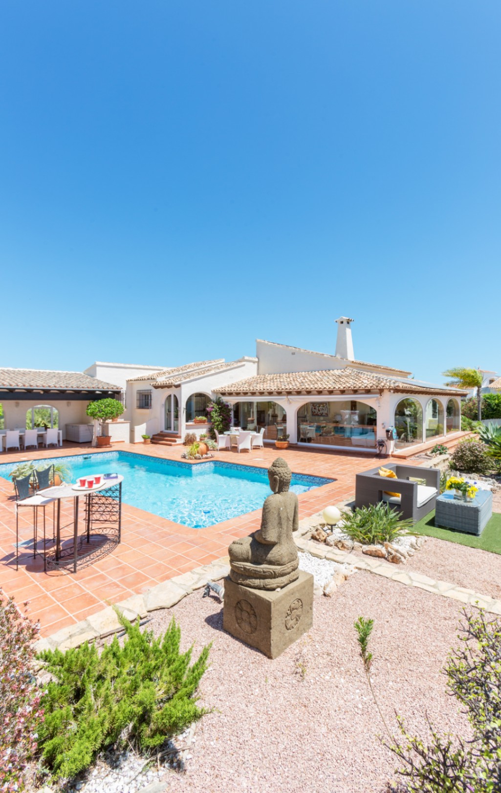 Außenbereich mit Pool, Terrakotta-Fliesen, einer Buddha-Statue aus Stein und einer mediterranen Villa mit verglastem Verandabereich.