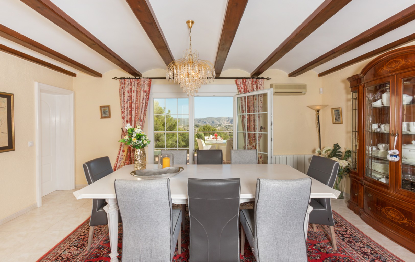 Esszimmer mit sichtbaren Holzbalken, weißem Esstisch und klassischem Vitrinenschrank. Große Glastüren führen zur Terrasse mit Blick auf die umliegende Landschaft.