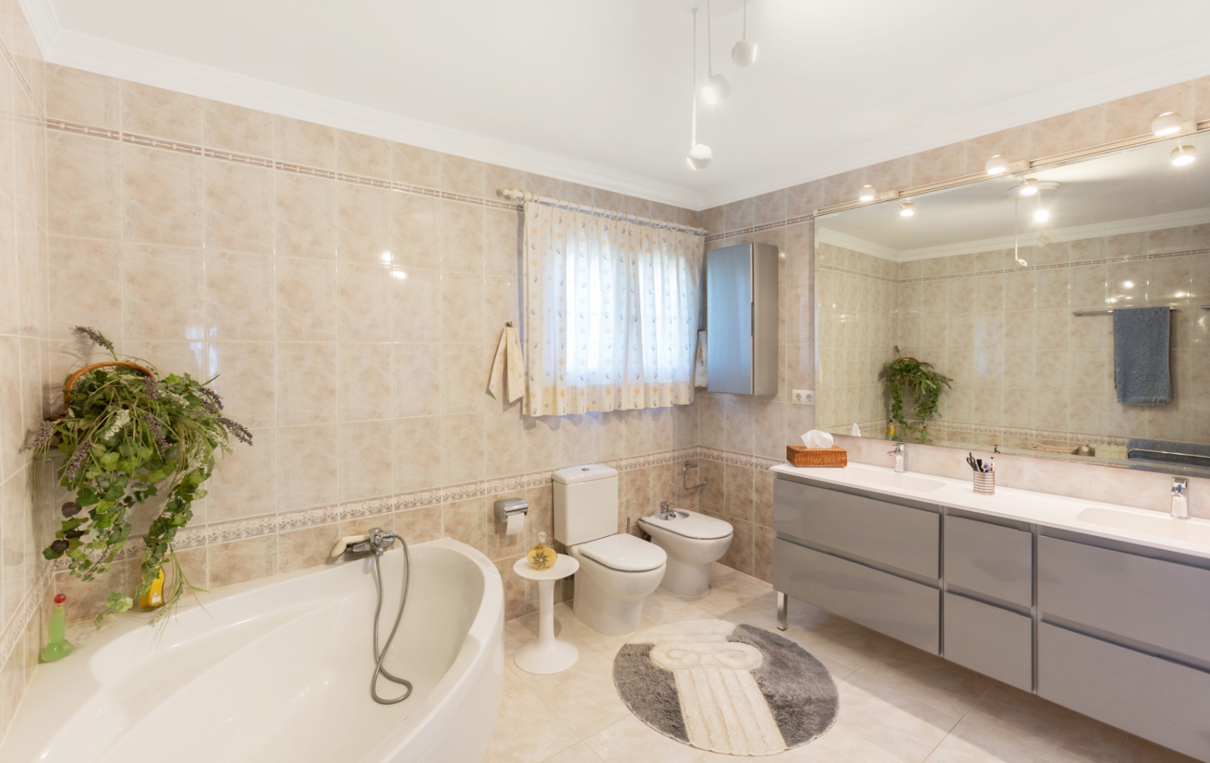 Geräumiges Badezimmer mit Eckbadewanne, Doppelwaschtisch in Grau und großem Wandspiegel. Beige Fliesen mit Zierbordüren an den Wänden und am Boden.
