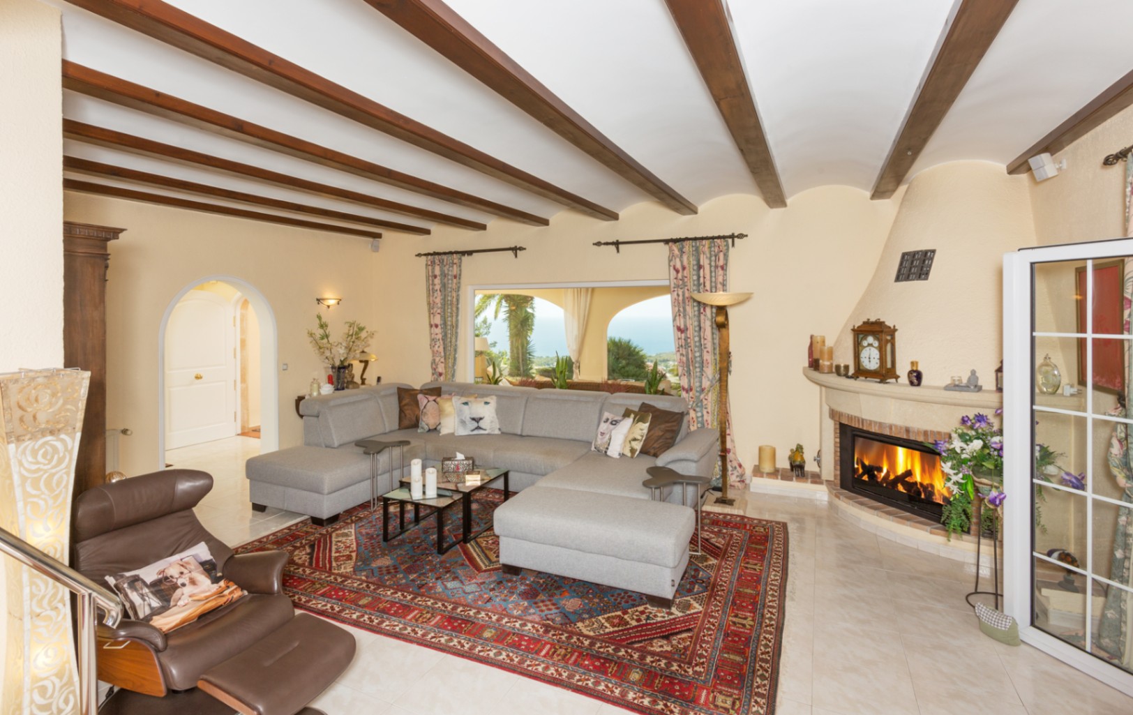Großzügiges Wohnzimmer mit sichtbaren Deckenbalken aus Holz, grauem Ecksofa, gemauertem Kamin und Meerblick durch das Panoramafenster.