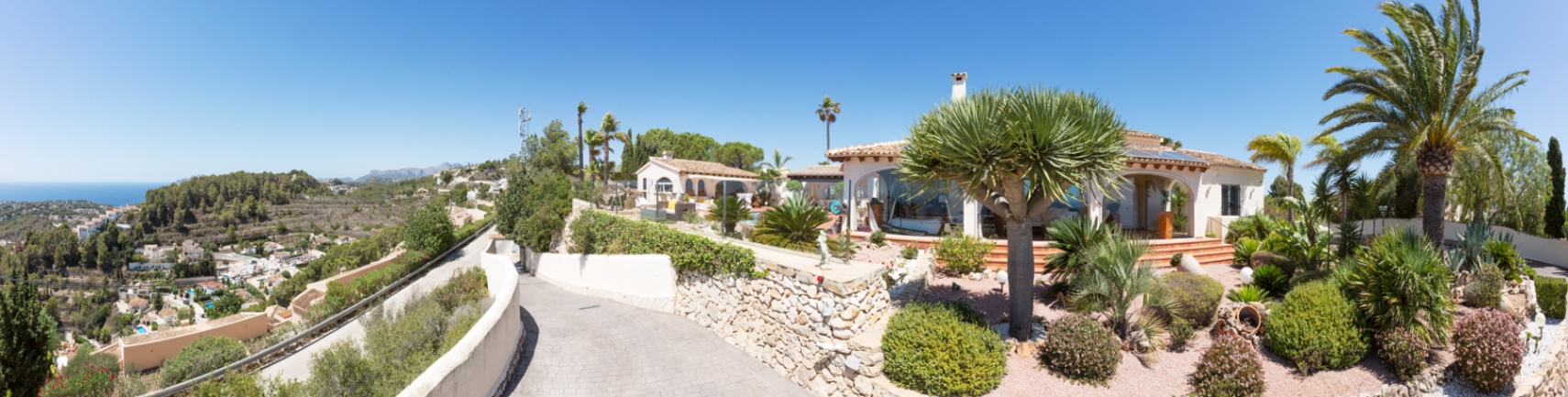 Panorama-Außenansicht der Villa mit gepflasterter Einfahrt, Trockensteinmauern und einem mediterran angelegten Garten mit Palmen und Blick auf die Küste.