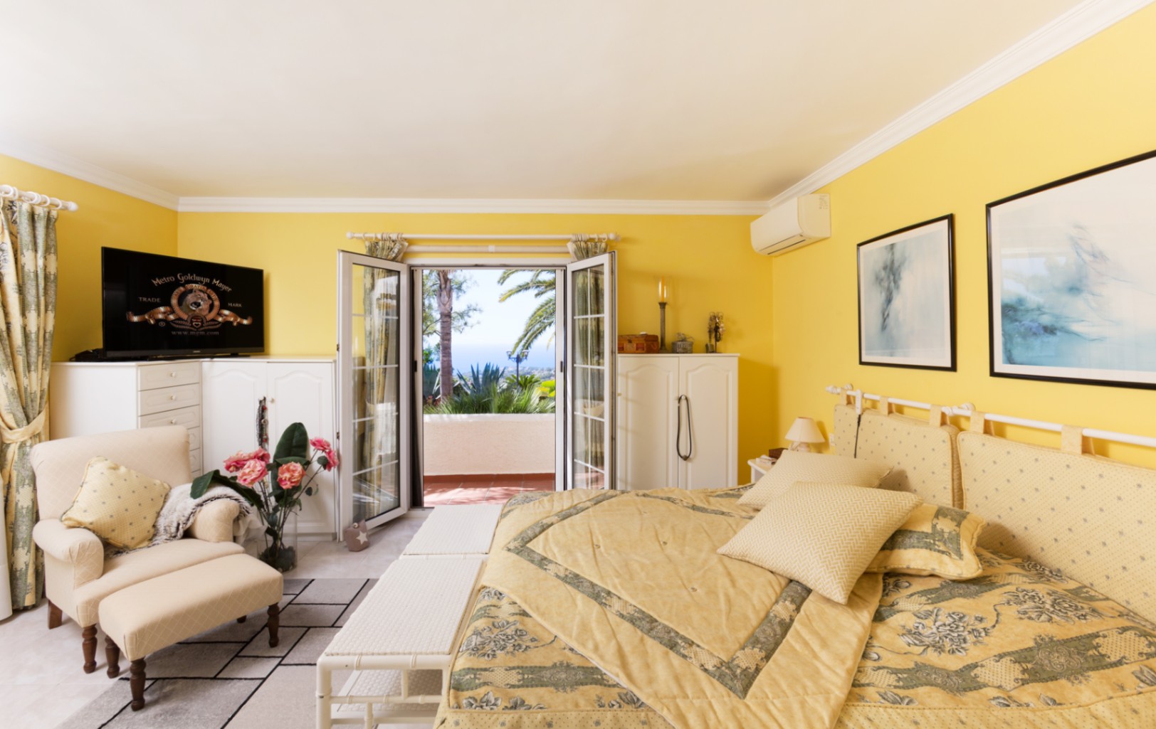 Schlafzimmer mit gelben Wänden, weißen Schränken und Sitzecke. Glastüren führen auf eine Terrasse mit Blick auf das Meer, Palmen und mediterrane Pflanzen.