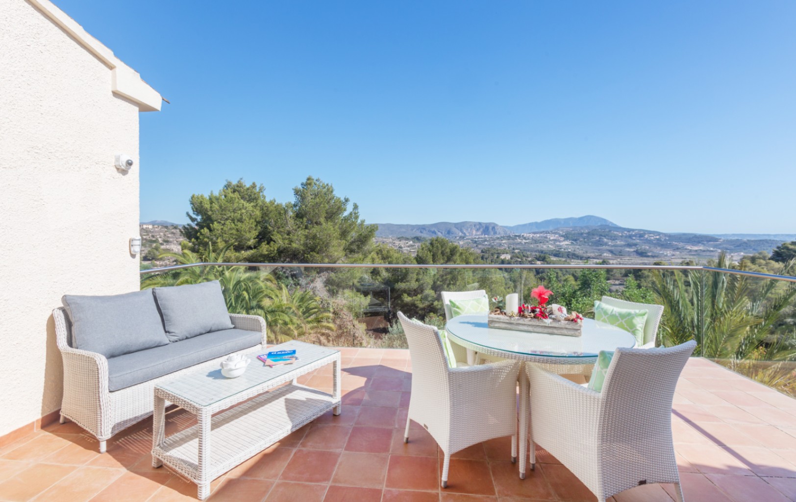 Terrasse mit Terrakotta-Fliesen, weißen Rattanmöbeln und Glasgeländer, die einen weiten Blick auf die umliegende Berglandschaft und Pinienwälder bietet.