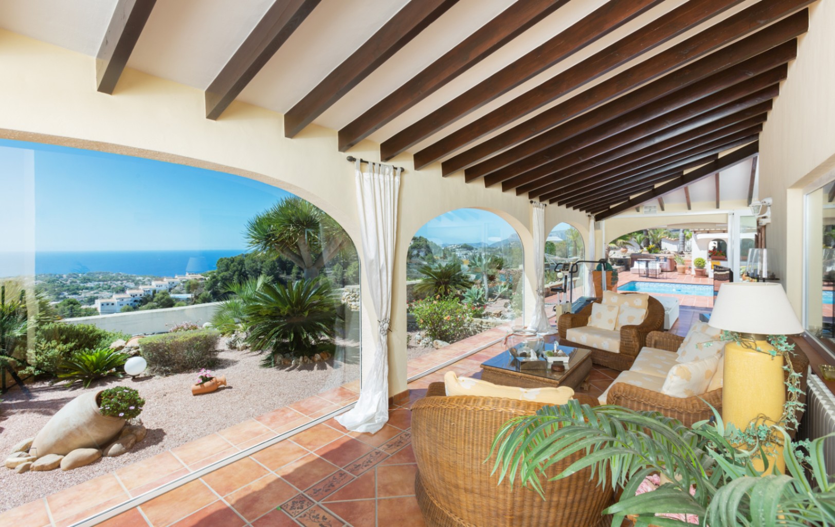 Überdachte Terrasse mit dunklen Holzbalkendecken, Terrakottafliesen und Korbmöbeln. Verglaste Rundbögen eröffnen einen Panoramablick auf das Meer und den Garten.