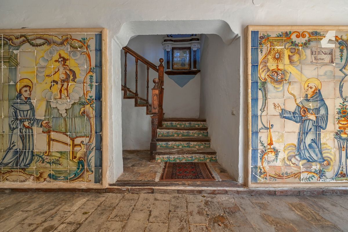 Eingangsbereich mit historischen, handbemalten Keramikwandbildern, weiß getünchten Steinwänden und einer Treppe mit dekorierten Setzstufen und Holzgeländer.