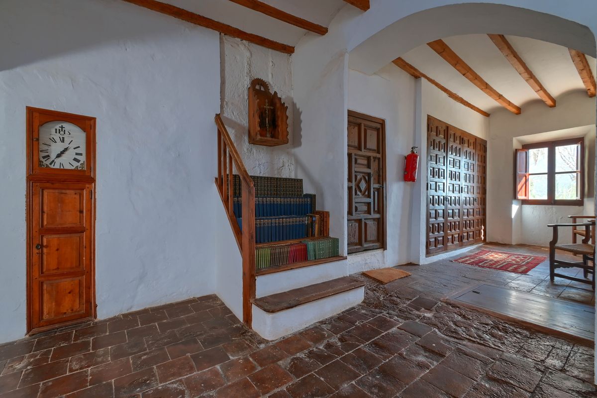 Flur mit Terrakottafliesen, Holzbalken und eingebauter Standuhr. Eine kleine Treppe mit integrierten Bücherregalen steht neben traditionellen Holztüren.