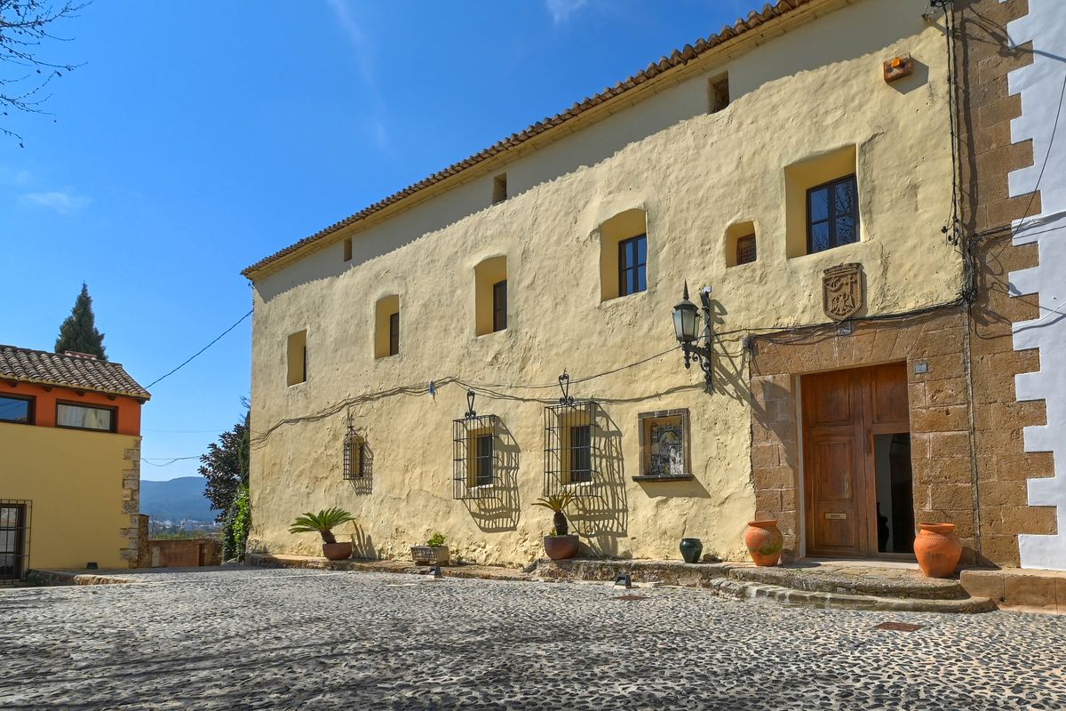 Historische Fassade mit gelben Putzwänden, einem Portal aus Tosca-Stein und Wappenrelief. Der Vorplatz ist mit traditionellem Kopfsteinpflaster und Terrakotta-Töpfen gestaltet.