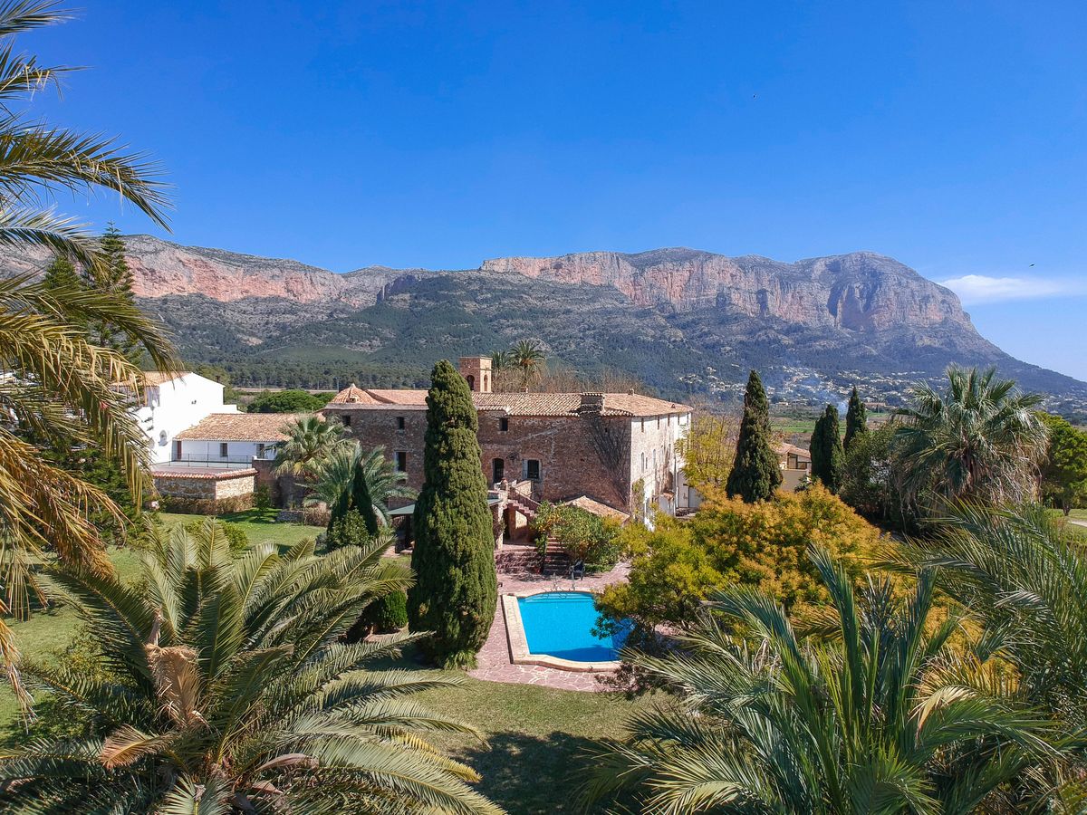 Historische Stein-Finca mit Swimmingpool, üppigen mediterranen Gärten und Panoramablick auf das Montgó-Massiv unter blauem Himmel.
