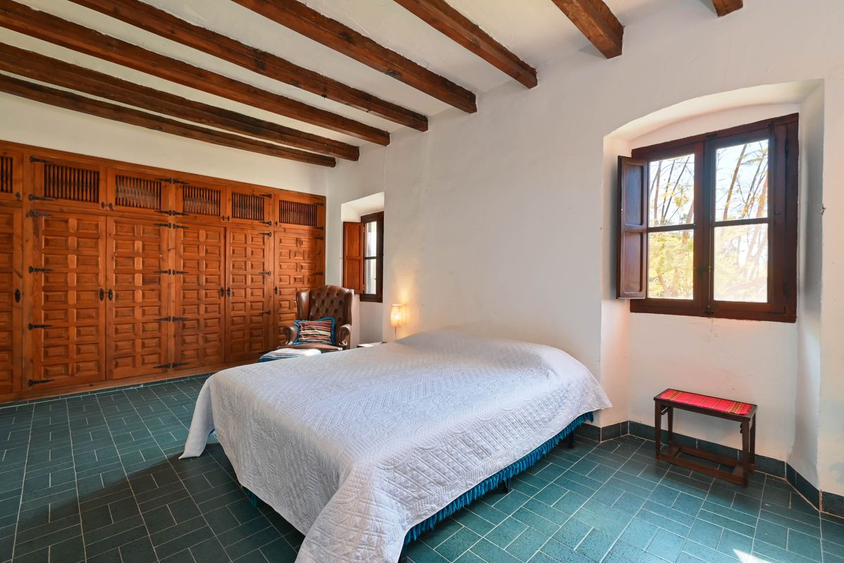 Rustikales Schlafzimmer mit sichtbaren Holzbalkendecken, Einbauschränken aus geschnitztem Holz, grünem Fliesenboden und tiefen Fenstern mit Fensterläden.