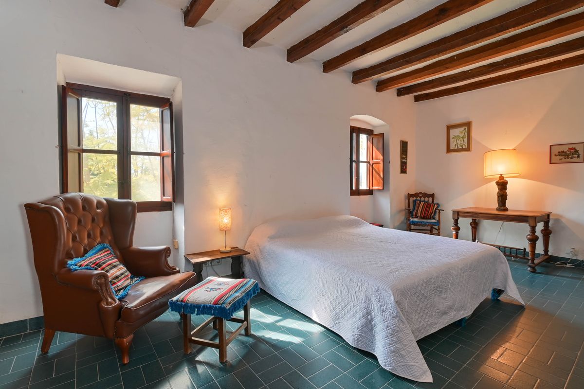 Rustikales Schlafzimmer mit sichtbaren Holzbalkendecken, grünen Fliesenböden und einem Leder-Ohrensessel neben traditionellen Holzfenstern mit Fensterläden.