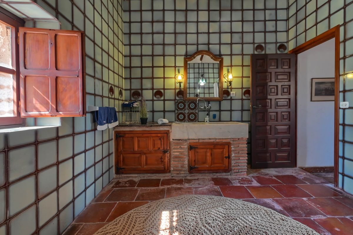 Traditionelles Badezimmer mit Steinwaschbecken auf Backsteinsockel, rustikalen Holzschränken, Terrakottaboden und markanten quadratischen Wandfliesen.