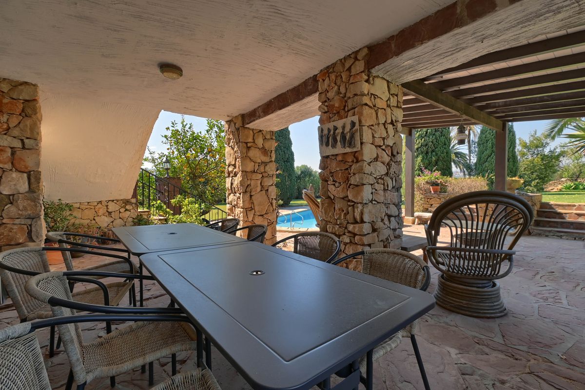 Überdachte Terrasse mit massiven Natursteinpfeilern, Außenessbereich und Blick auf den Pool sowie die angrenzende Gartenanlage.
