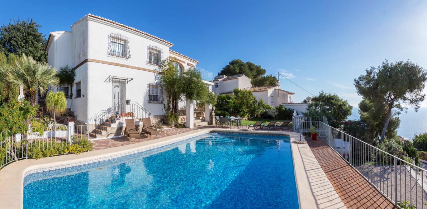 Außenansicht der mediterranen Villa mit großem Swimmingpool, Naturstein-Terrasse, Sonnenliegen und weißer Fassade mit traditionellen Fenstergittern und Meerblick.