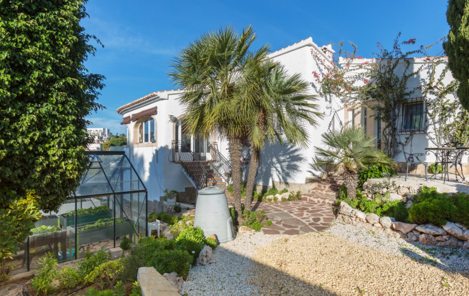 Außenansicht einer weißen mediterranen Villa mit Natursteinwegen, Palmen und einem Gewächshaus inmitten einer gepflegten Gartenanlage.