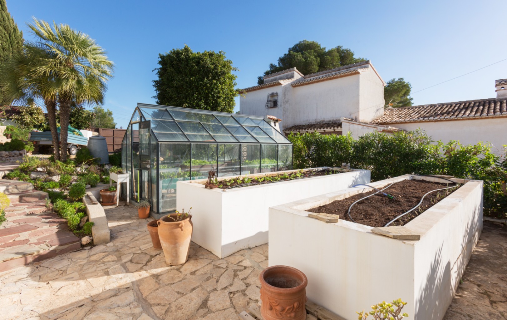 Gartenbereich mit einem Gewächshaus aus Glas und zwei großen, weiß verputzten Hochbeeten mit integrierter Bewässerung auf einer Naturstein-Terrasse.